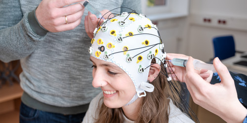 Eine Probandin trägt die EEG-Kappe mit den Kabeln und Elektroden. Die Studiendurchführer*innen passen die Kappe an.