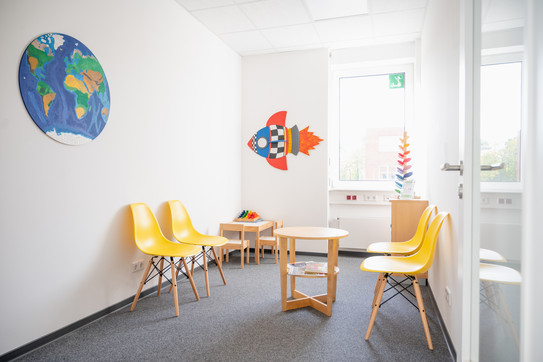 Der Raum in dem die Proband*innen warten ist bunt gestaltet. Die Plastikstühle sind gelb und der Boden besteht aus Teppich. Ein großes Fenster gibt natürliches Licht und an der Wand steht ein Regal mit verschiedenen Spielen für die Kinder. An der Wand hängen eine aufgemalte Erdkugel und eine Rakete.