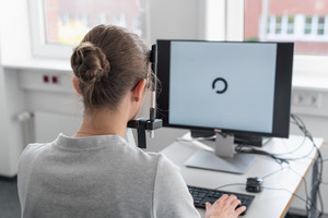 Eine Probandin sitzt an einem Schreibtisch mit dem Testungscomputer, dessen Bildschirm einen Kreis abbildet, der zu einer Seite eine Öffnung aufweist.