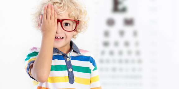 Junge hält sich eine Augenseite zu und trägt eine Brille. Im Hintergrund ist ein Sehtest, welcher verschwommen ist.