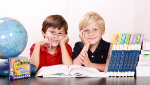 zwei Jungs lehnen stützen ihr Kinn auf ihren Händen ab und stützen die Ellenbogen auf dem Tisch auf. Auf dem Tisch stehen ein Globus und mehrere Lehrbücher. Ein Lehrbuch liegt aufgeschlagen vor ihnen.