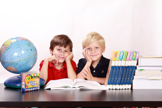 zwei Jungs lehnen stützen ihr Kinn auf ihren Händen ab und stützen die Ellenbogen auf dem Tisch auf. Auf dem Tisch stehen ein Globus und mehrere Lehrbücher. Ein Lehrbuch liegt aufgeschlagen vor ihnen.