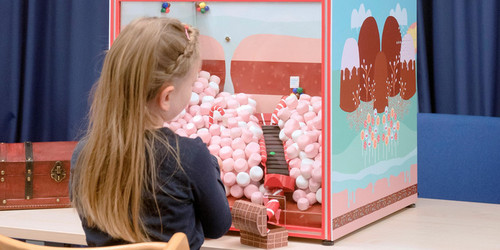 Kind sitzt mit Rücken zur Kamera und blickt auf eine pinke Spielbox in etwa der Größe eines Kleinkindes. In der Box sind pinke watteähnliche Kügelchen und zwischen ihnen führt eine Treppe entlang. Der Hintergrund ist mit pinken Hügeln bemalt.