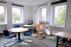 Der Diagnostikraum mit zwei Tischen (rund und eckig) und Stühlen am runden Tisch. im Hintergrund steht ein Schreibtisch mit einem Computer und daneben eine Box für eine Studie. Der Raum ist mit vier großen Fenstern begrenzt.