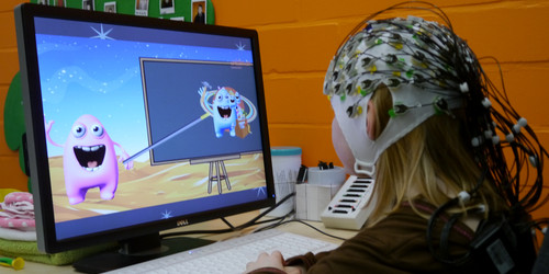 Kind sitzt mit EEG-Kappe am Computer auf dem ein spielerischer Bildschirm abgebildet ist