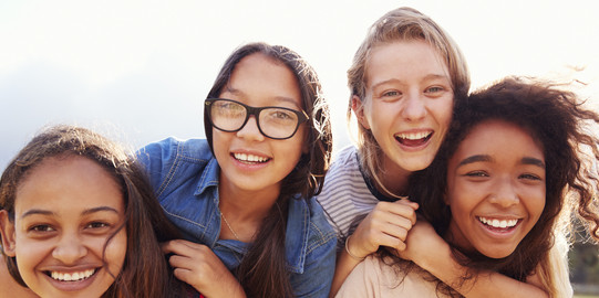 vier Mädchen lachen in die Kamera. Zwei der Mädchen tragen die beiden anderen Mädchen auf ihrem Rücken. Ein Mädchen trägt eine Brille.