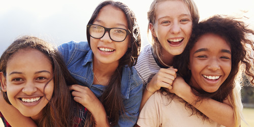 vier Mädchen lachen in die Kamera. Zwei der Mädchen tragen die beiden anderen Mädchen auf ihrem Rücken. Ein Mädchen trägt eine Brille.