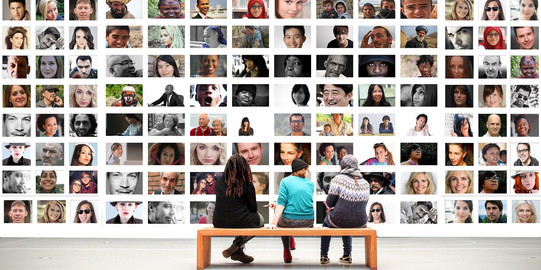 Viele Fotos von Gesichtern erschrecken sich über das gesamte Bild und drei Personen sitzen vor dieser Bilderwand und betrachten sie