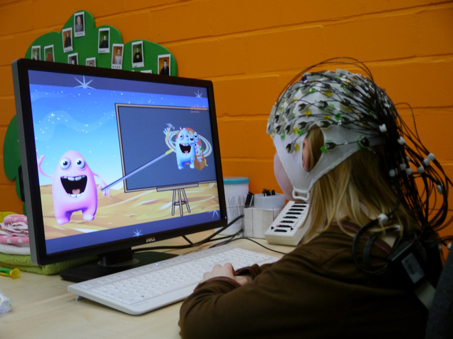 Kind sitzt mit EEG-Kappe am Computer auf dem ein spielerischer Bildschirm abgebildet ist