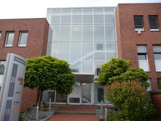 Das Gebäude in der Otto-Hahn-Straße 20 von außen. Dier Flur ist verglast und es befinden sich kleine Bäume vor dem Eingang