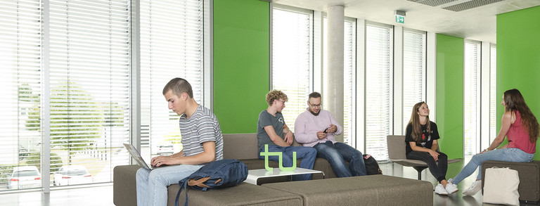 Sechs Studierende sitzen in einem Aufenthaltsbereich in einem Uni-Gebäude. Sie lesen Bücher, arbeiten am Laptop oder unterhalten sich.